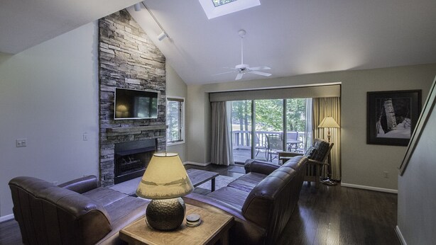 A living room with a stone fireplace, a ceiling fan, and a view of the outdoors.