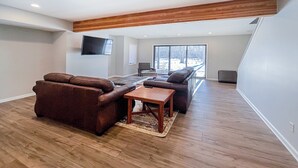 A spacious living room with a wooden ceiling, a large window, and a flat-screen TV mounted on the wall.