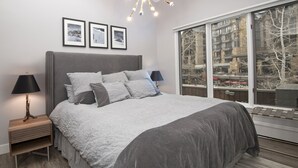 Een moderne slaapkamer met een groot bed, twee nachtkastjes met lampen, een kroonluchter, ingelijste foto’s aan de muur en een groot raam met uitzicht op gebouwen.