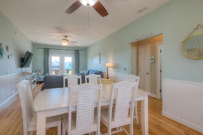 Un comedor con una mesa y sillas, un ventilador de techo, un televisor sobre un mueble bajo y una puerta con la leyenda 'Cafe Island'.