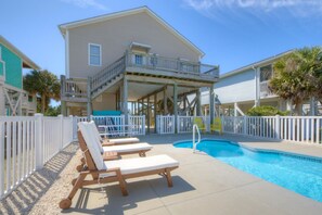 A pool area with lounge chairs, a covered deck, and a two-story building with a balcony. - A World Away: spacious 4BR beach home, perfect for 8 guests. (Oak Island)