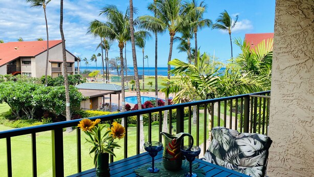 A balcony with a view of a swimming pool, palm trees, and a building with a red roof. - Deluxe Two Bedroom Patial Ocean View Luana Kai Resort B308 (Kihei)