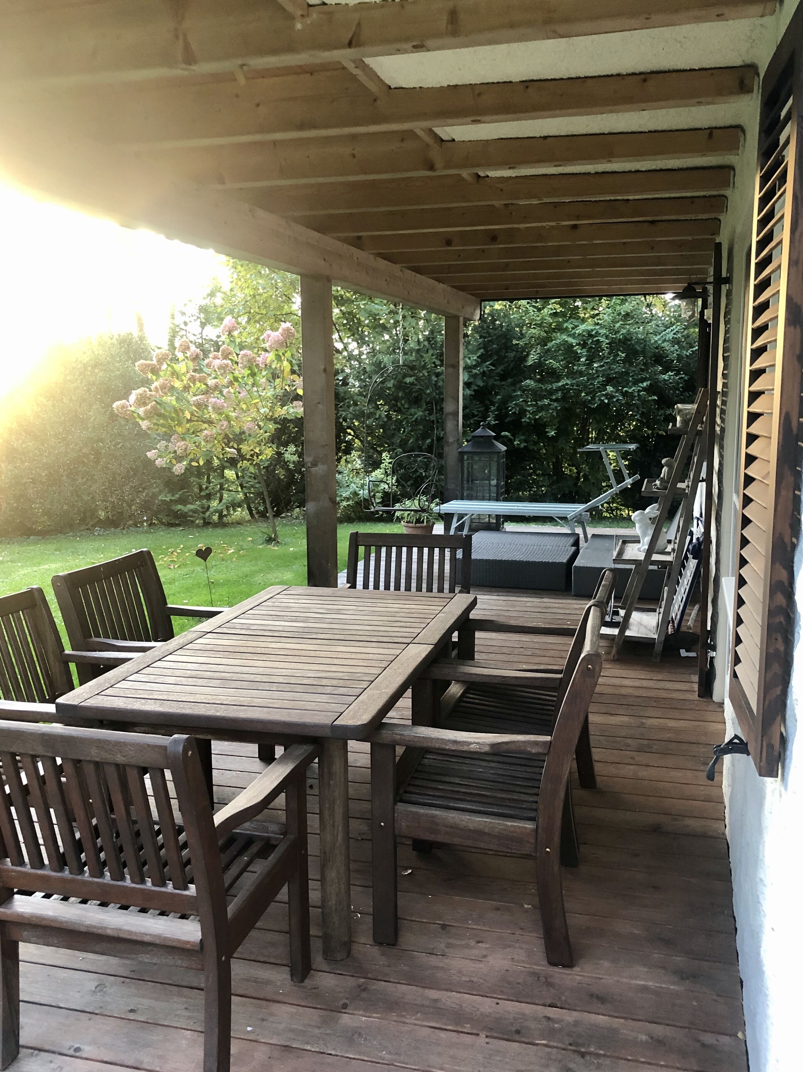 Un patio techado al aire libre con mobiliario de madera, vista a un jardín y una estructura de pérgola.