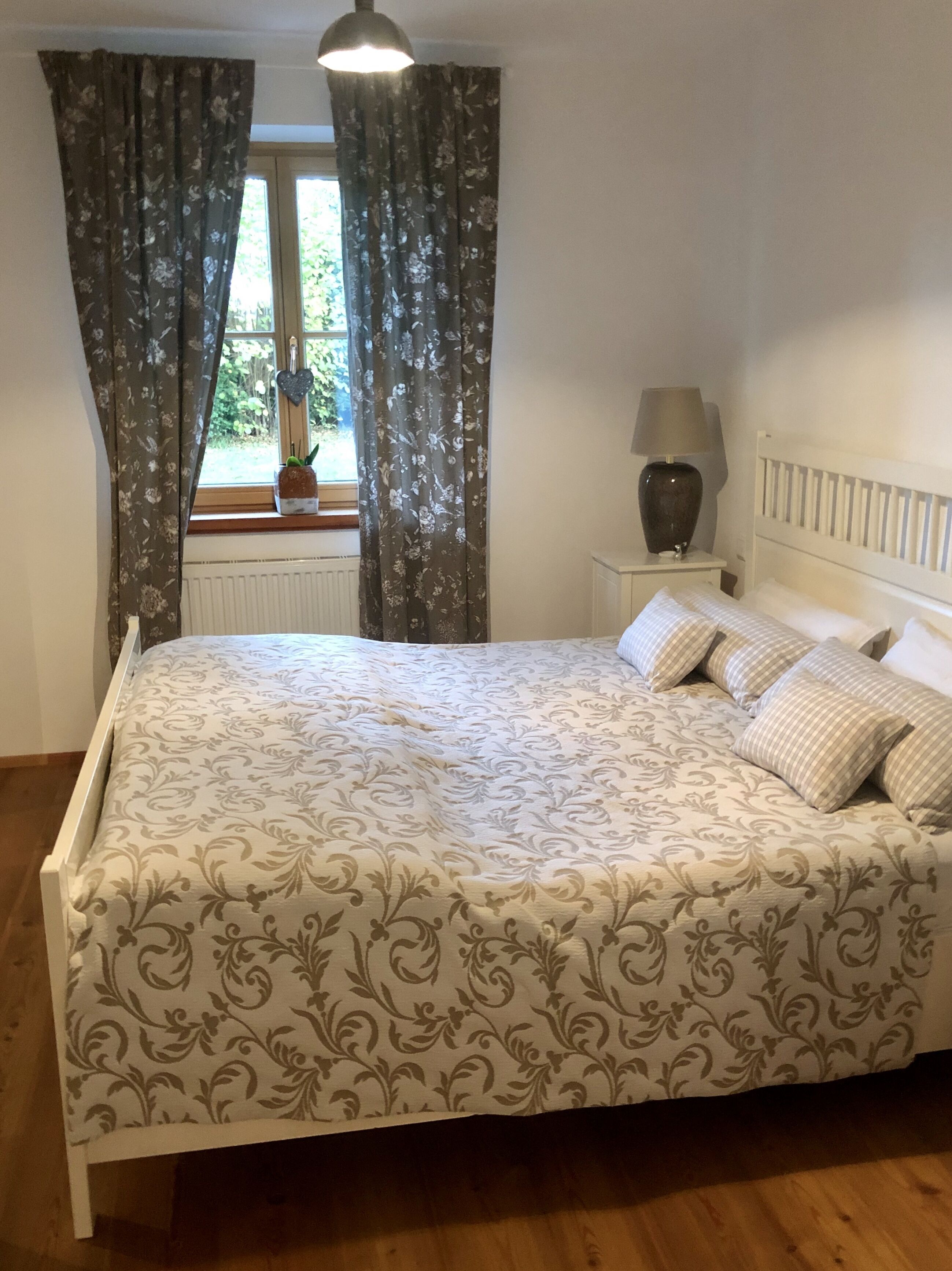 A bedroom with a bed, curtains, a nightstand, a lamp, and a window with a view of greenery.