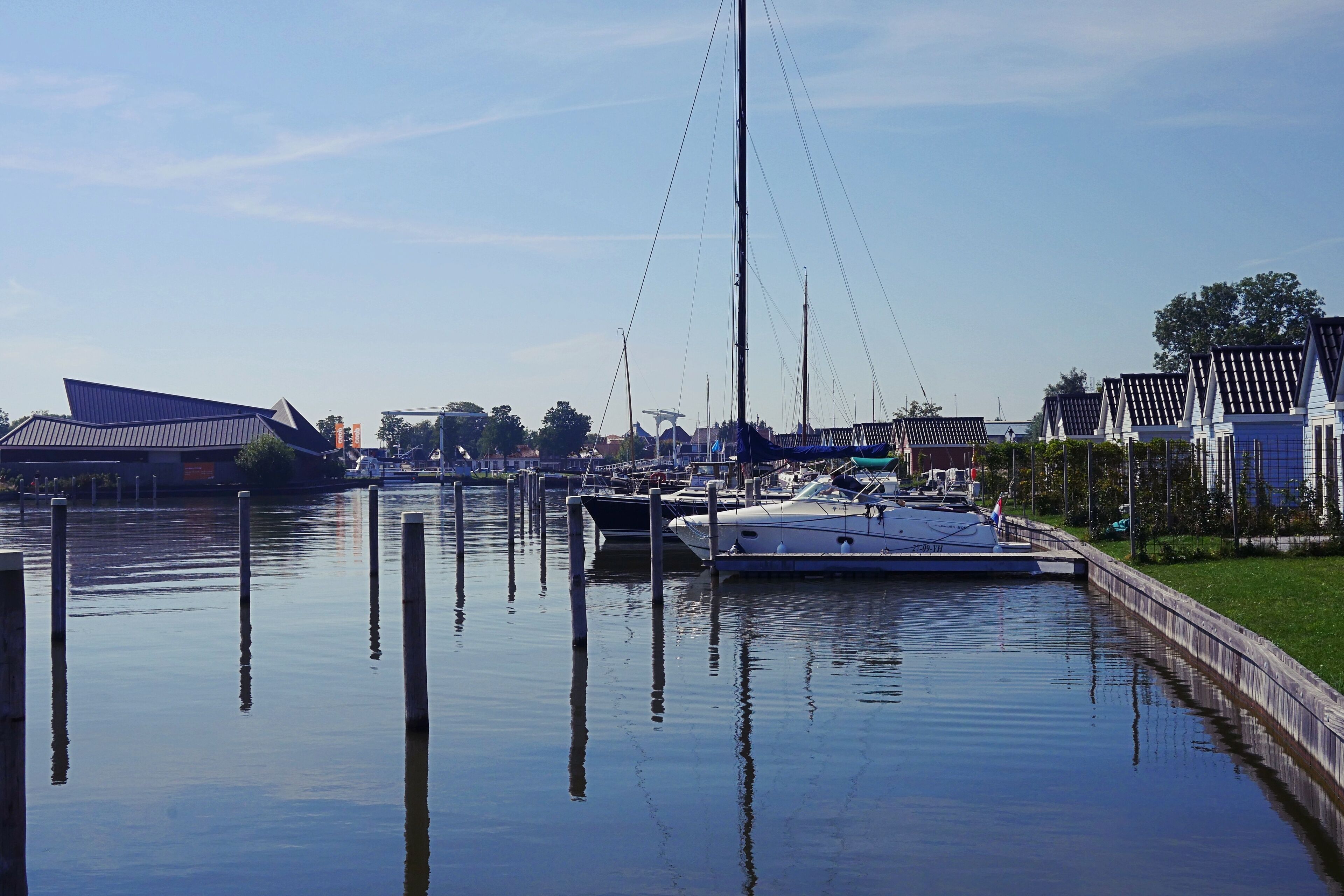 Een jachthaven met aangemeerde boten, een brug op de achtergrond en een modern gebouw met een schuin dak.