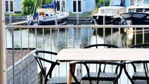 Blick auf einen Hafen mit vertäuten Booten, eine Holzterrasse mit Stühlen und Häuser im Hintergrund.