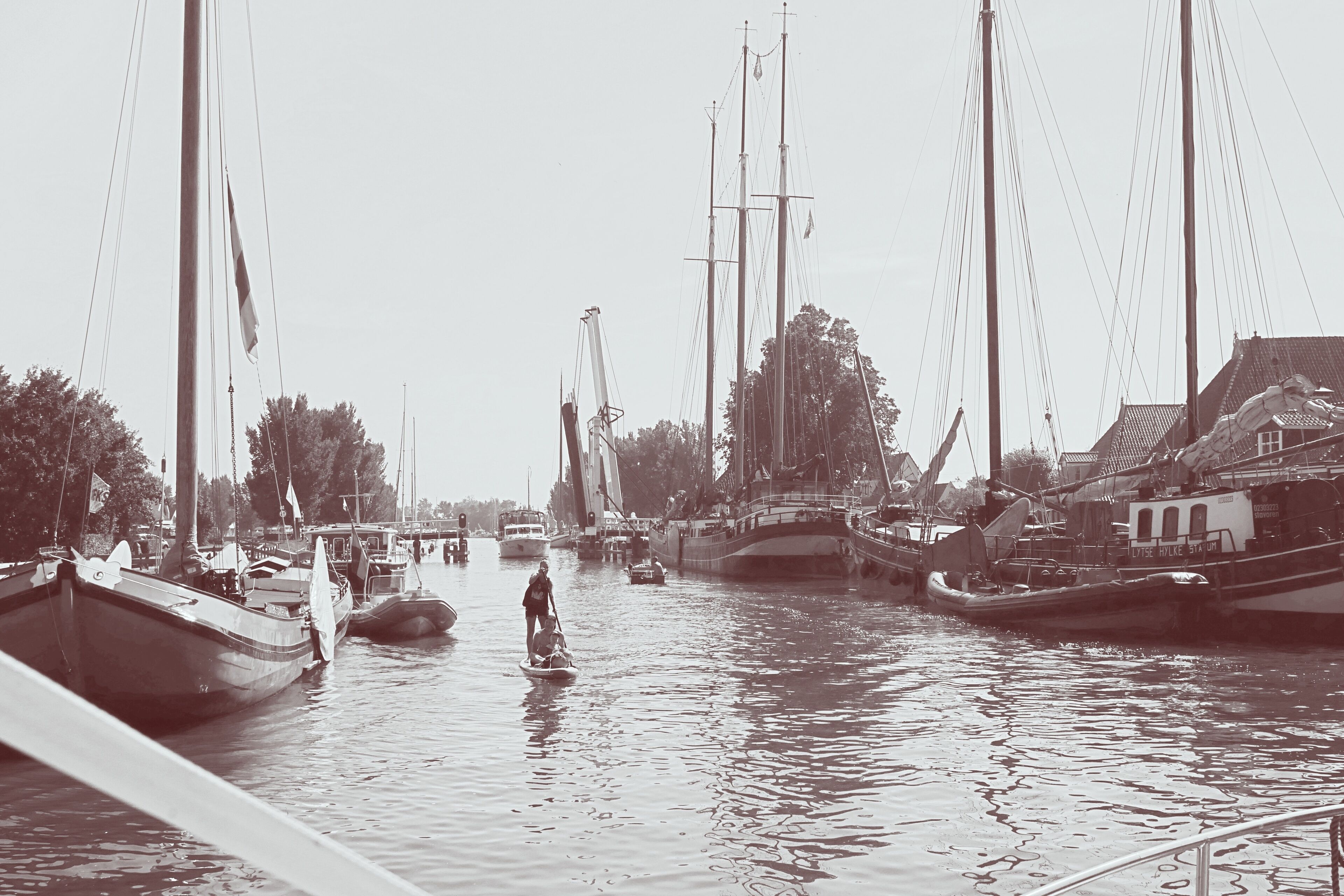 Un port où plusieurs bateaux sont amarrés, une personne sur une planche à pagaie et des bâtiments qui bordent la rive.