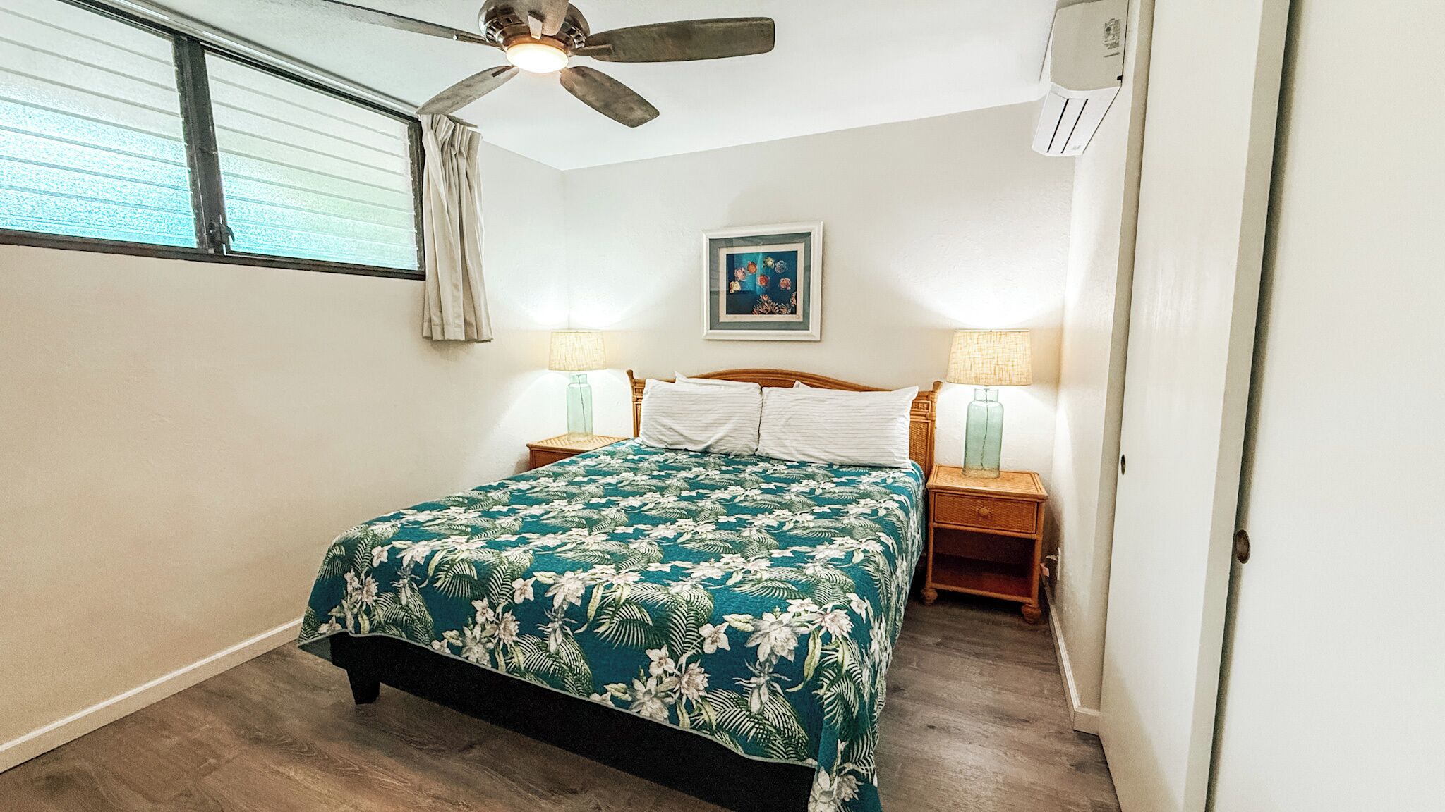 Une chambre à coucher comprenant un lit, deux tables de chevet, un ventilateur de plafond, une fenêtre avec stores et un climatiseur fixé au mur.