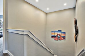 A hallway with a white wall, a set of stairs, and a framed picture of a sunset over a canyon.
