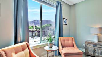 Una habitación con un ventanal grande, dos sillones, una mesita con una planta y un armario de madera con una lámpara.