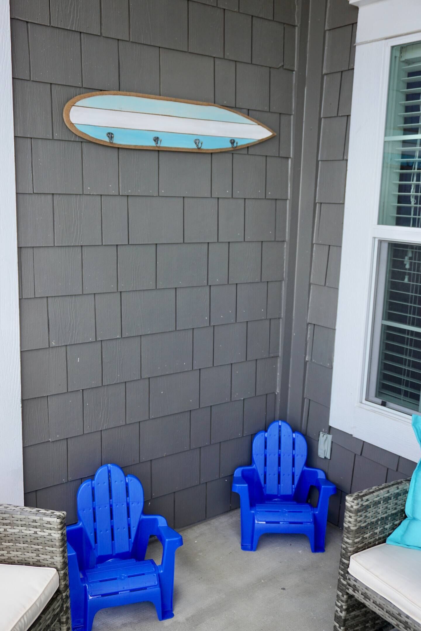 Una esquina con dos sillas Adirondack azules, un adorno en forma de tabla de surf y una ventana con persianas.