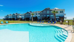 Une piscine entourée de chaises bleues, un immeuble doté de nombreux balcons et un ciel d’un bleu limpide.
