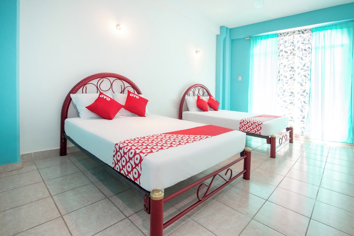 Dos camas con ropa de cama roja y blanca, una pared azul y una ventana con cortinas traslúcidas.
