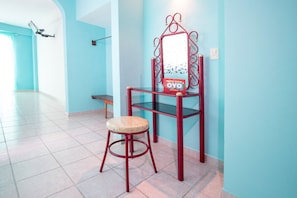 A room with a stool, a red chair with a mirror, and a wall-mounted shelf.