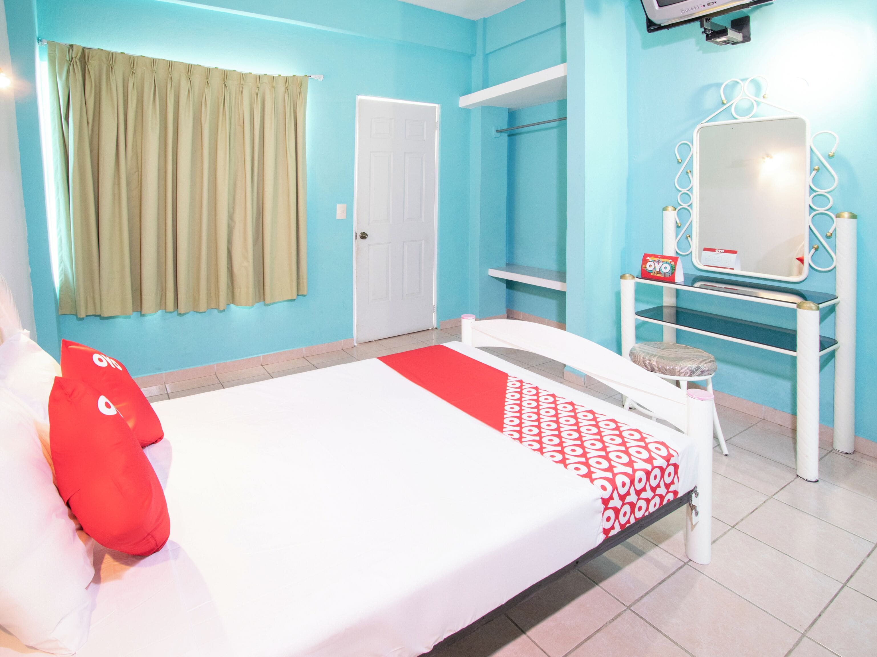 Una habitación de hotel con una cama, un tocador con espejo, una puerta y una estantería con un objeto rojo.