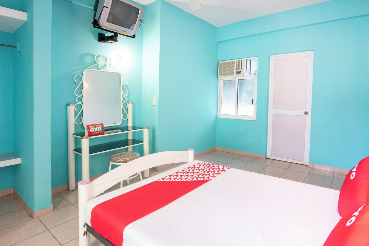Una habitación con una cama, un televisor montado en la pared, una ventana y un equipo de aire acondicionado, además de una puerta.