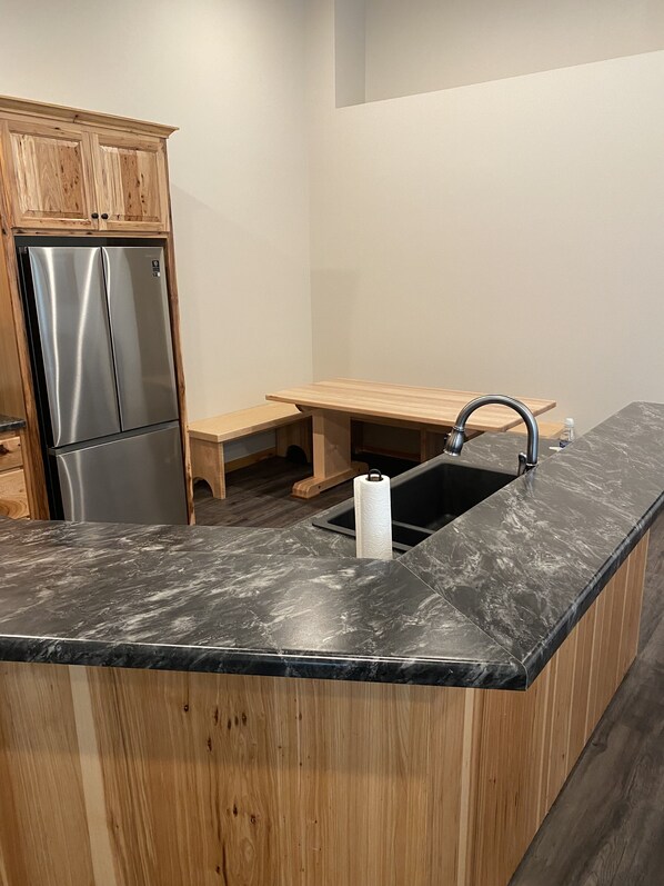 A kitchen with a marble countertop, stainless steel refrigerator, wooden cabinets, and a wooden bench.