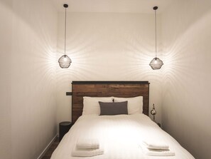 Un lit bien fait, avec du linge de lit blanc et deux serviettes pliées au pied du lit, une tête de lit en bois et deux lampes suspendues au motif tissé.
