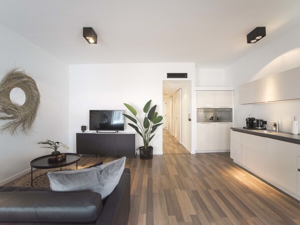 Un salon moderne avec un canapé, un téléviseur, une petite table et une plante en pot. Il y a également un coin cuisine avec des placards et un plan de travail.