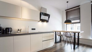 A modern kitchen with white cabinets, a wooden dining table with chairs, and a window with blinds. - Sack-Time 6: Luxury & relaxation near the sea! (Zandvoort)