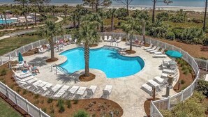 Una piscina circular con sillas de descanso y palmeras, rodeada por una valla, con una playa y el océano al fondo.