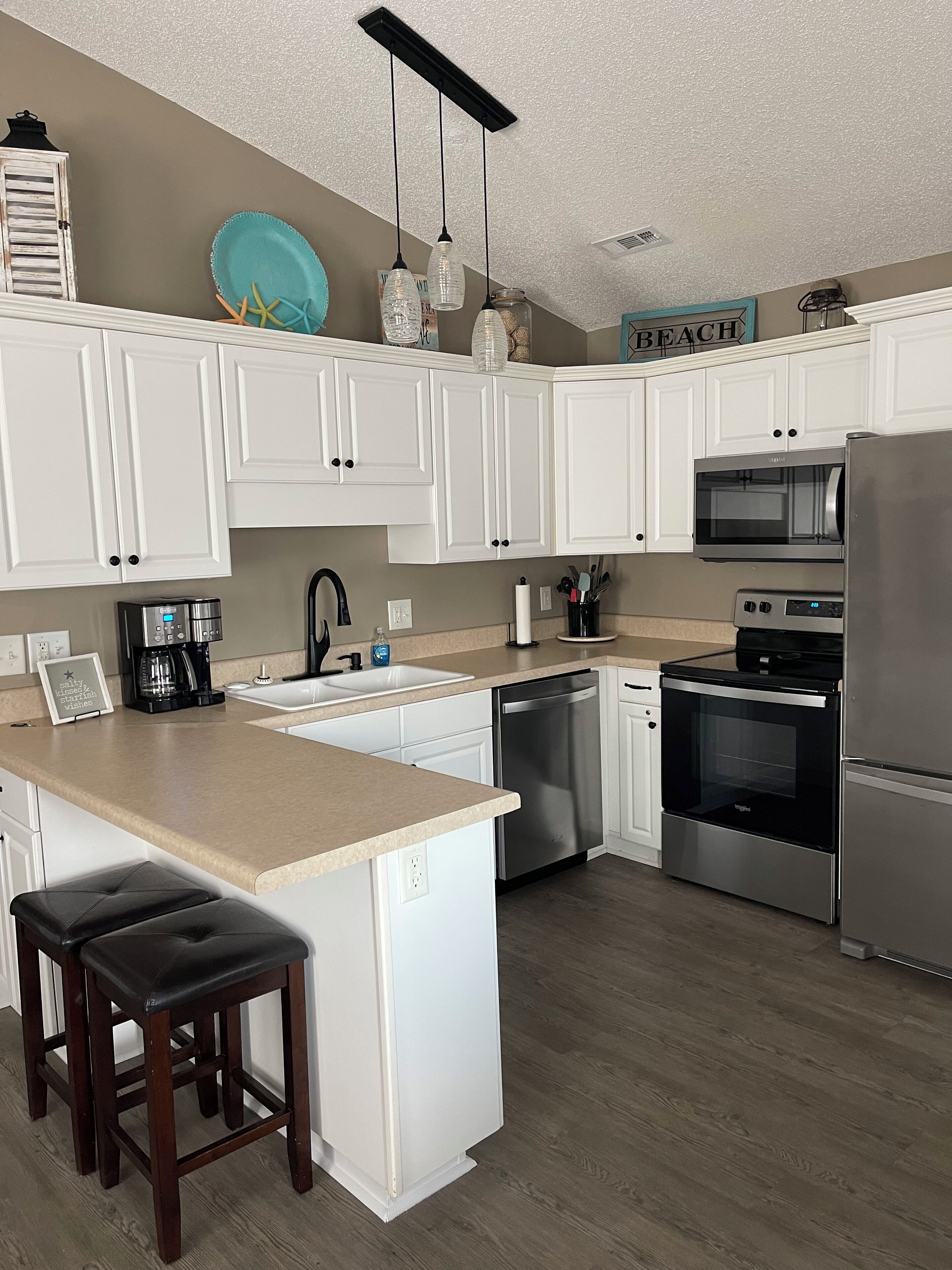 A modern kitchen with white cabinets, stainless steel appliances, a central island with stools, and a sign reading "BEACH".
