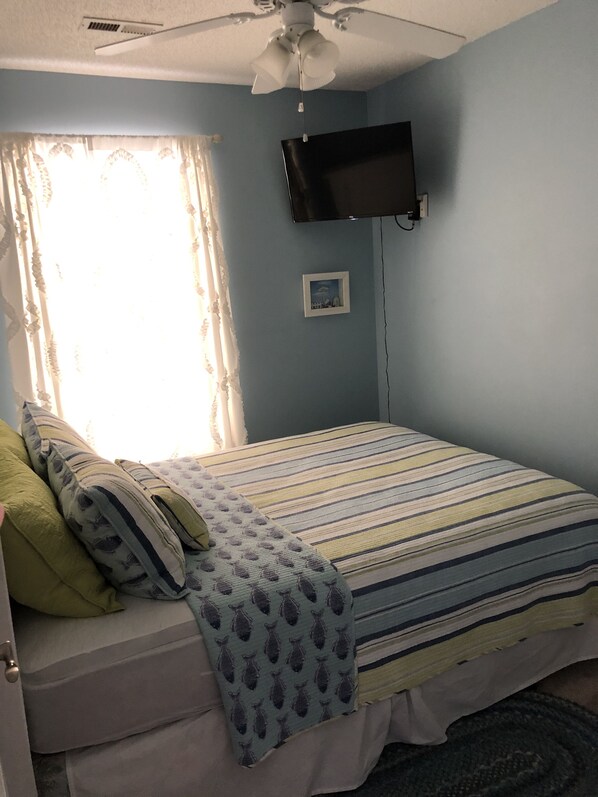 A bedroom with a bed, a ceiling fan, a flat-screen TV mounted on the wall, and a window with curtains.