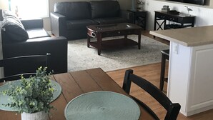 A living room with a sofa, coffee table, and dining table with placemats. There's a ceiling fan, a TV, and a painting on the wall. - Ocean Lakes  Clean Home for your Family! Now booking for 2026. (Myrtle Beach)