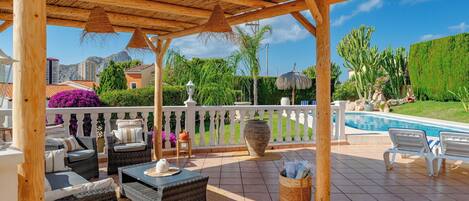 A covered outdoor seating area with wicker furniture, a large pot, and a pool in the background.