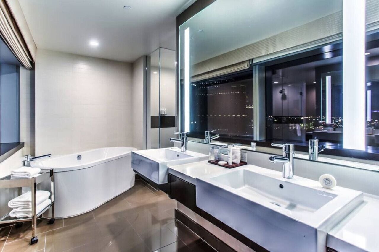A modern bathroom with two sinks, a large bathtub, and a mirror reflecting a cityscape.