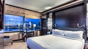 A modern hotel room with a large bed, a desk, and a view of a cityscape at night. - City Center! | Panoramic Corner Suite at Vdara (Las Vegas)