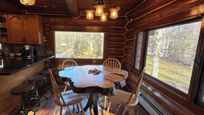 Interior rústico de una cabaña con una mesa de comedor de madera rodeada de sillas, una zona de cocina con armarios de madera y grandes ventanas que ofrecen vistas al bosque.