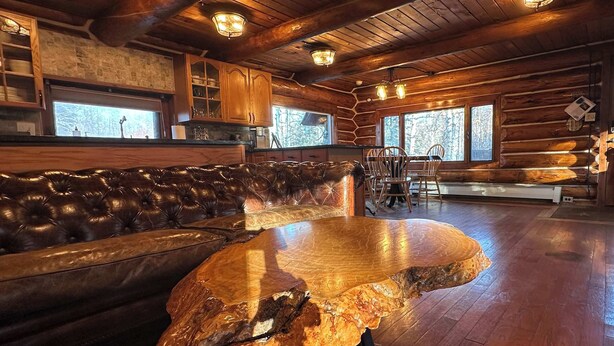 Interior de una cabaña de troncos con un banco de cuero, una mesa de madera de diseño único y un comedor con sillas y una ventana.