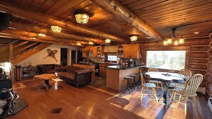 Un acogedor interior de cabaña con techo de madera, una zona de comedor con mesa y sillas, una cocina con armarios de madera y una sala de estar con sofá de cuero y chimenea.