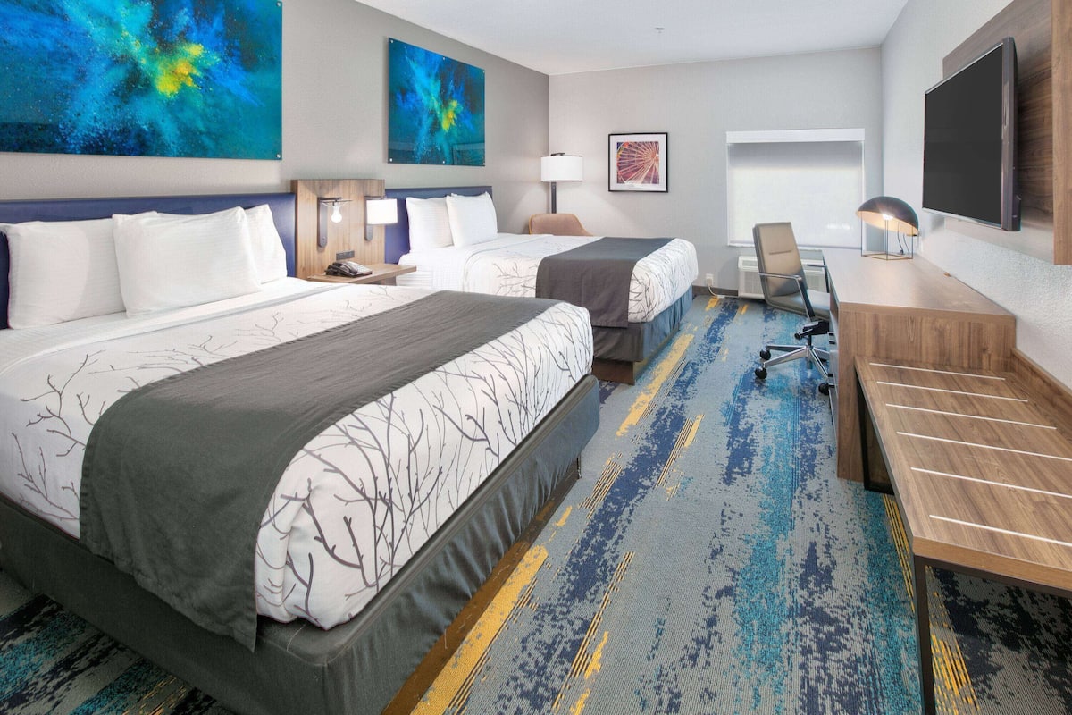 A hotel room with two beds, a desk, a chair, a TV, and artwork on the walls.
