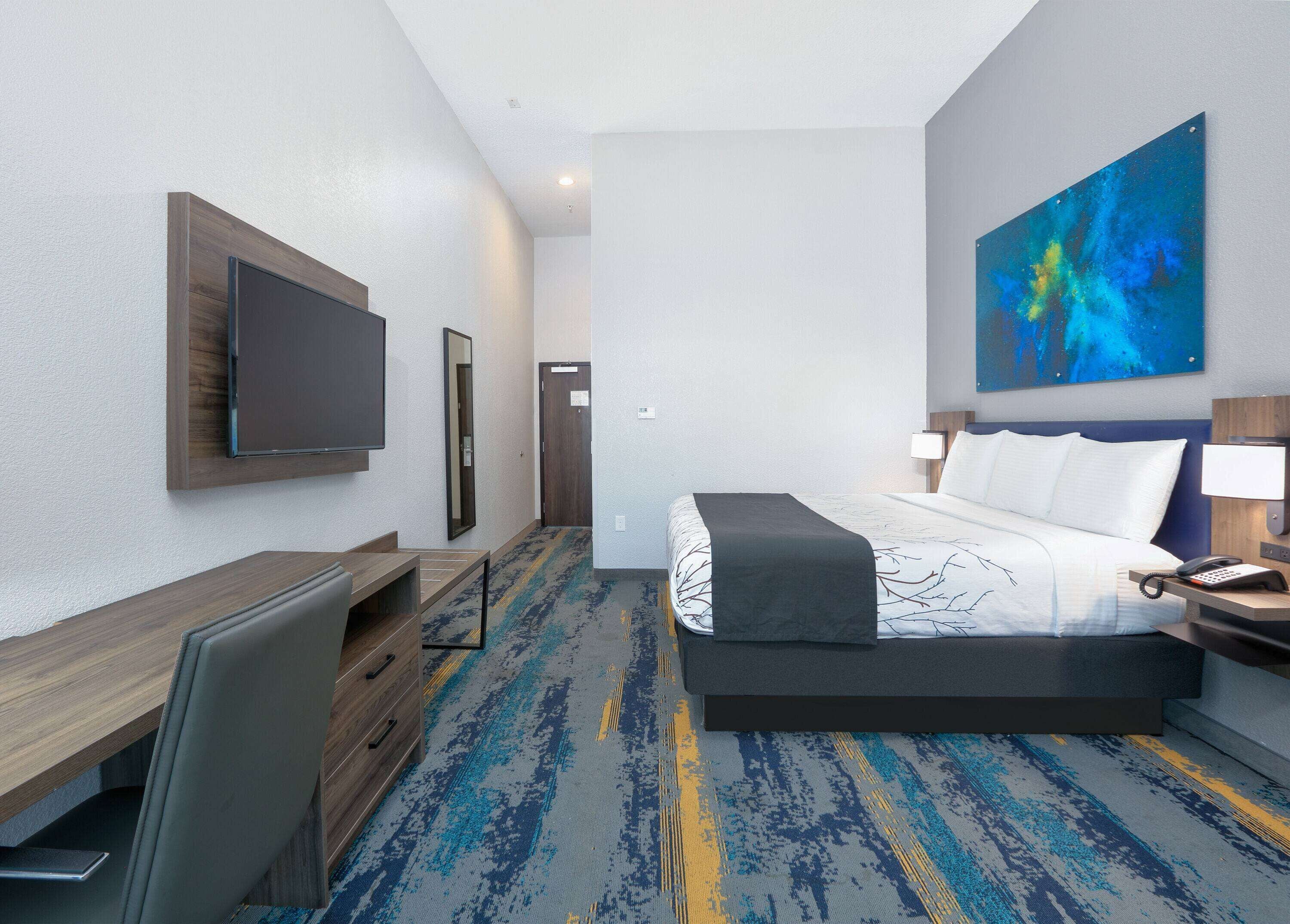 A hotel room with a bed, desk, chair, TV, and artwork on the wall.