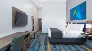 A hotel room with a bed, desk, chair, TV, and artwork on the wall.