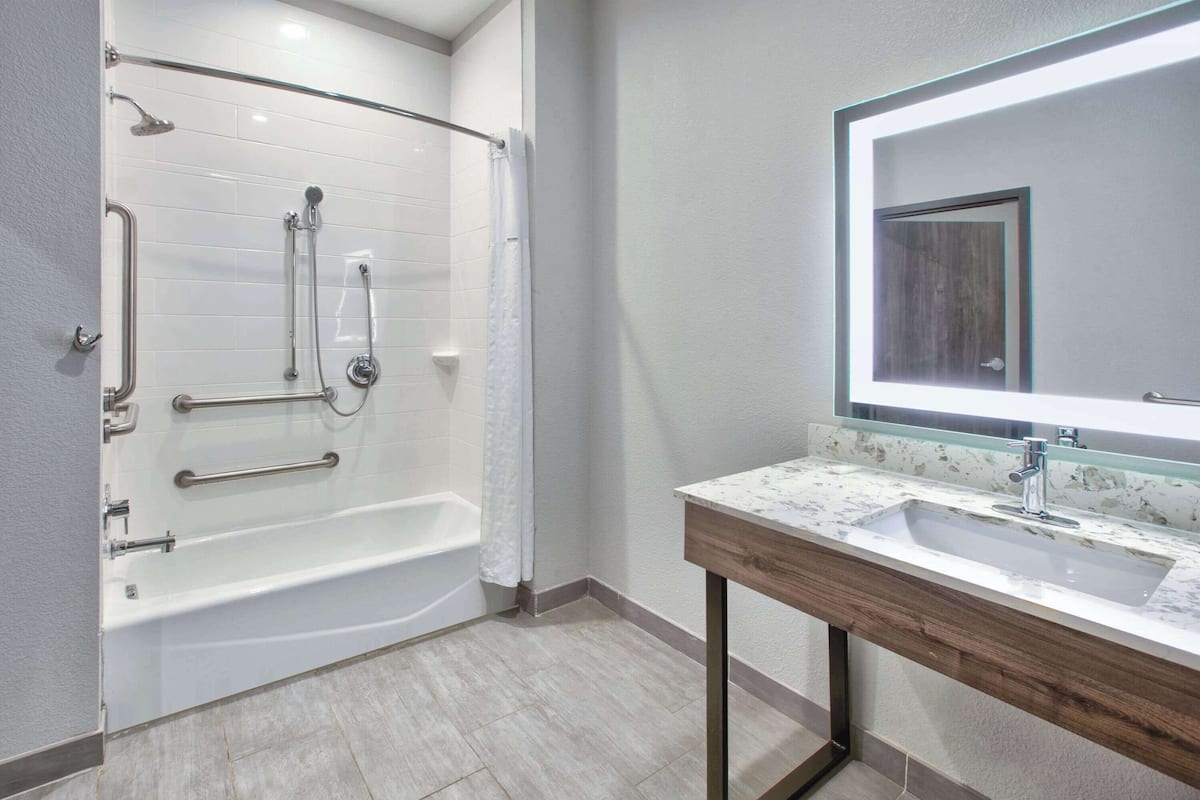 A modern bathroom with a walk-in shower, grab bars, a large mirror, and a marble countertop sink.