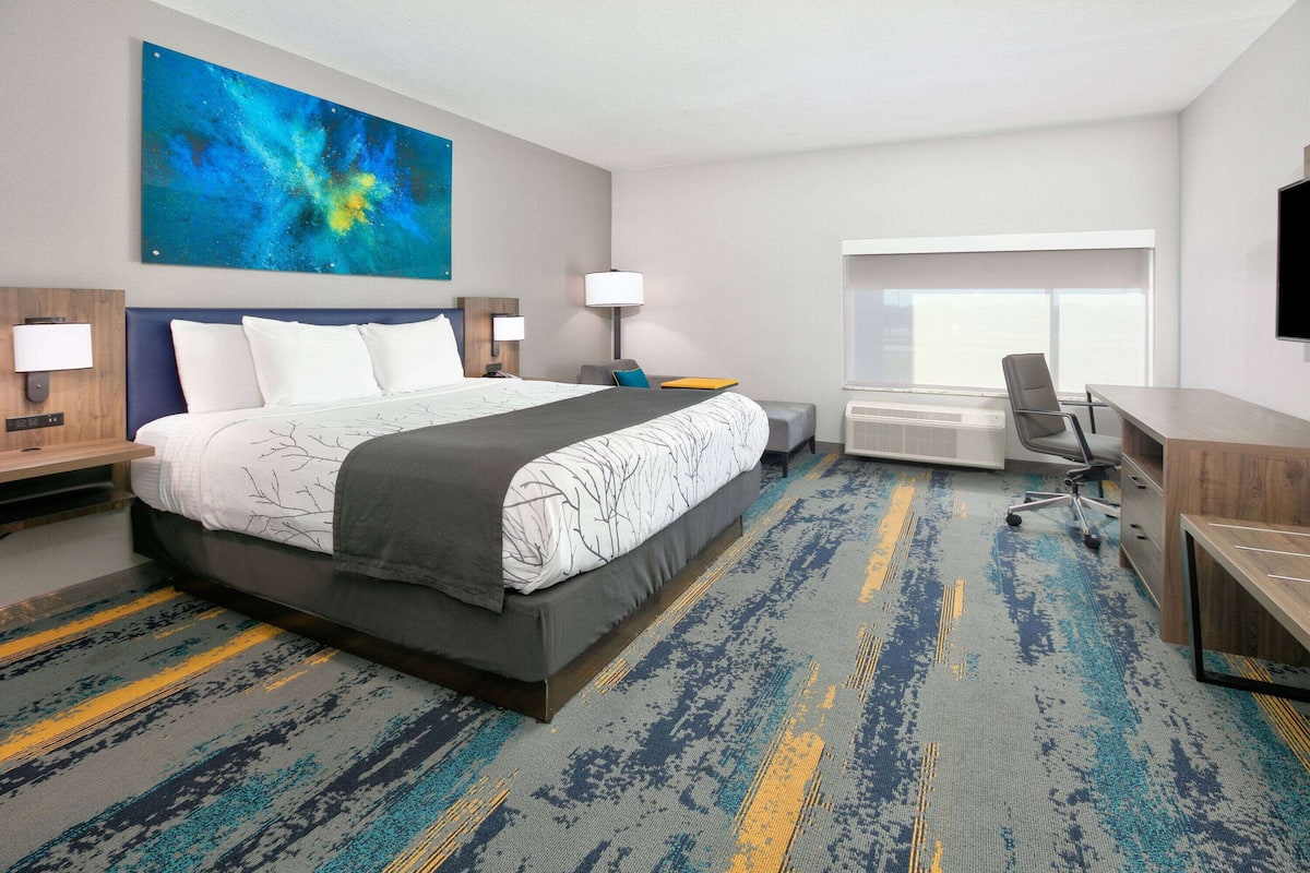 A hotel room with a large bed, a desk, a chair, a TV, and a large abstract painting on the wall.