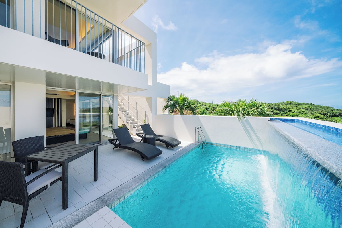 A modern pool area with a glass-enclosed upper level, a staircase, and a view of greenery.