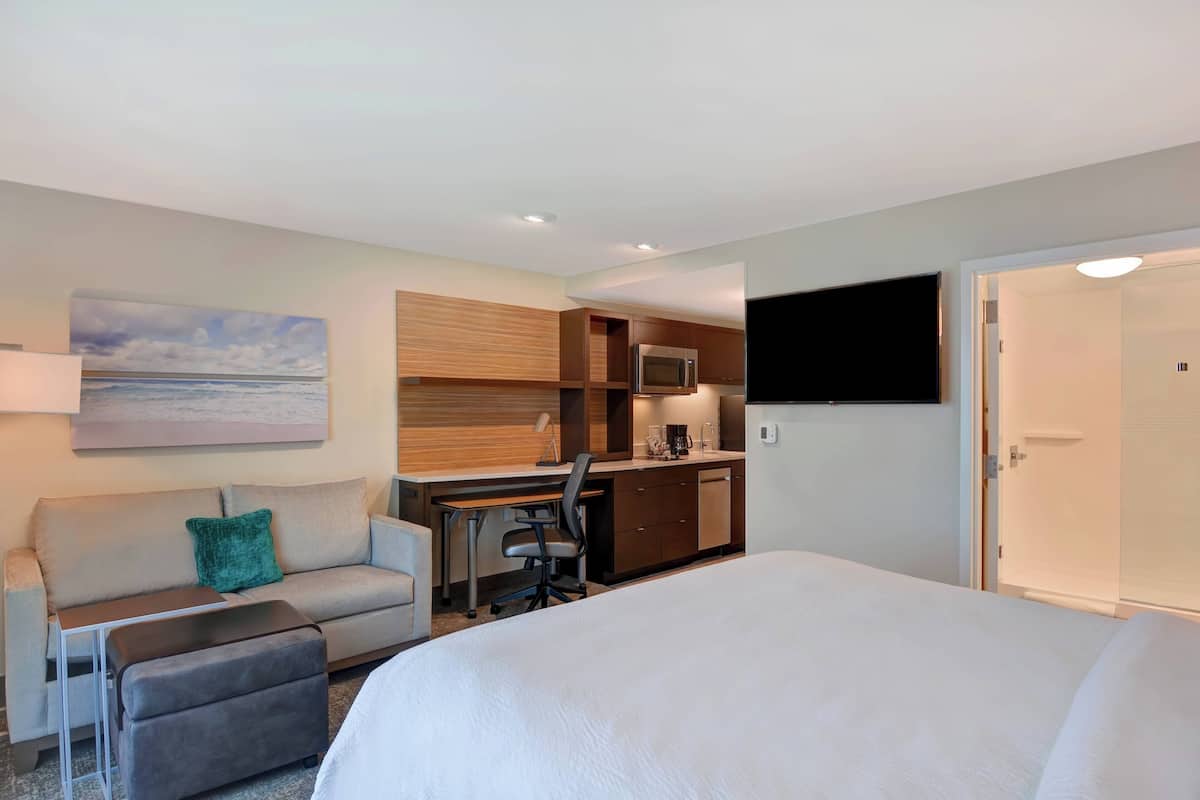 A hotel room with a bed, a desk with a chair, a kitchenette, a TV mounted on the wall, and a painting of a beach scene.