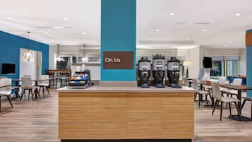 A modern coffee station with multiple coffee dispensers, a sign reading "On Us," and a seating area with tables and chairs.