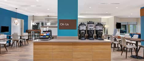 A modern coffee station with multiple coffee dispensers, a sign reading "On Us," and a seating area with tables and chairs.
