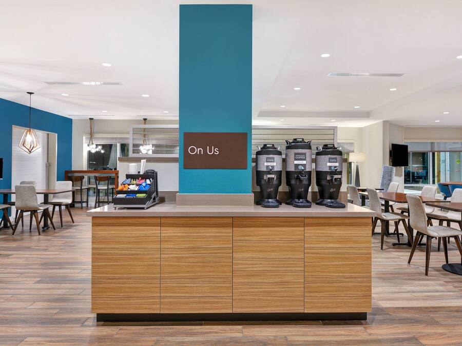 A modern coffee station with multiple coffee dispensers, a sign reading "On Us," and a seating area with tables and chairs.
