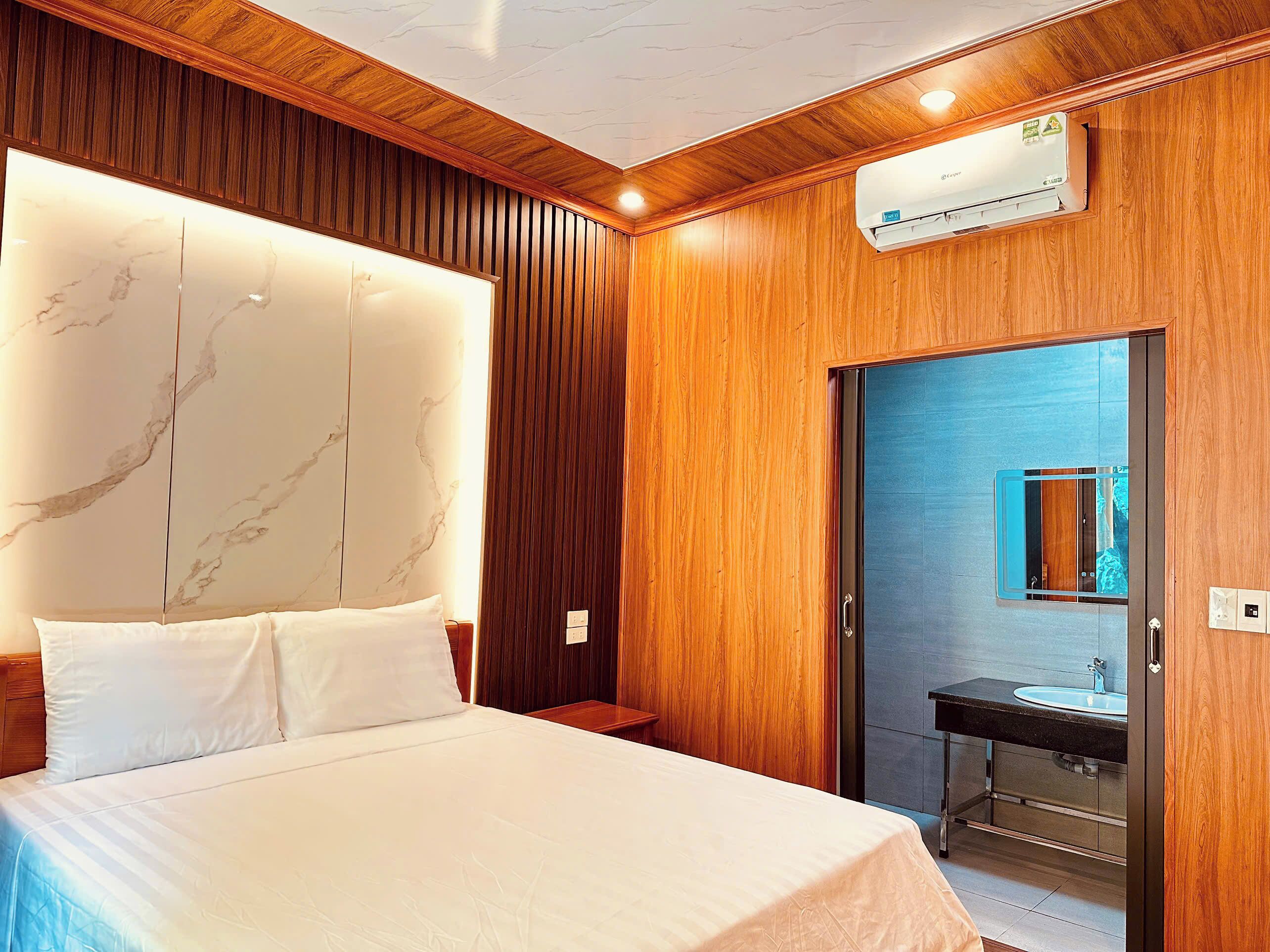 A hotel room with a bed, wooden panel walls, a marble headboard, a bathroom with a sink and mirror, and an air conditioning unit.