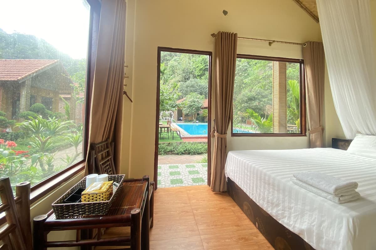 A bedroom with a bed, a window with curtains, a view of a pool and greenery, and a wooden chair with a basket.