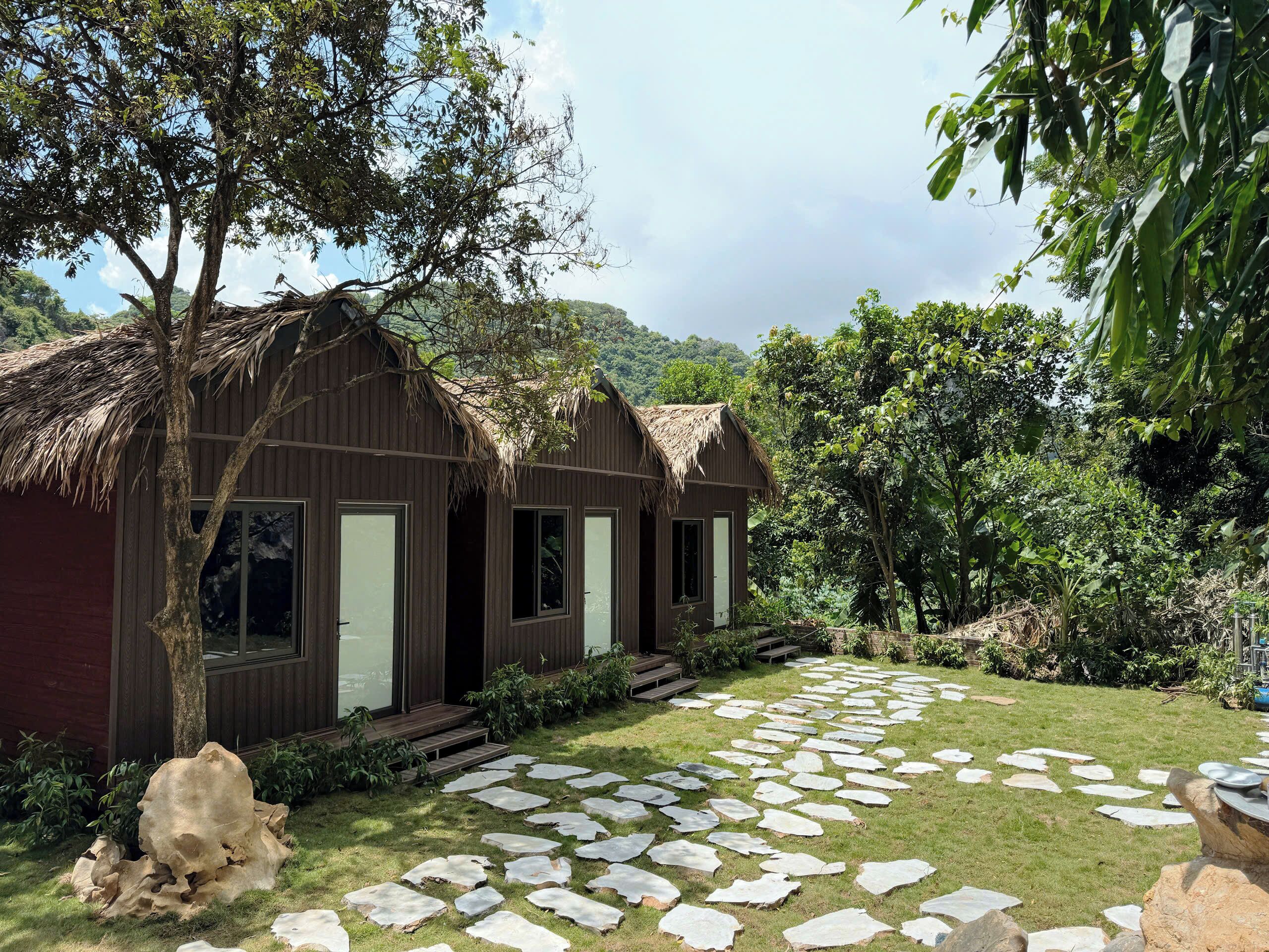 A rustic wooden building with a thatched roof, surrounded by lush greenery and a stone pathway.
