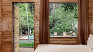 Een houten kamer met een bed, uitzicht op een zwembad en een weelderig groen landschap.