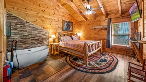 Une chambre de style rustique avec un plafond en bois, un plafond en pente, un grand lit, une baignoire, une fenêtre avec des stores et un téléviseur fixé au mur.