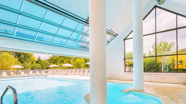 Una piscina interior con techos de cristal transparente, columnas blancas y vistas a zonas verdes al exterior.